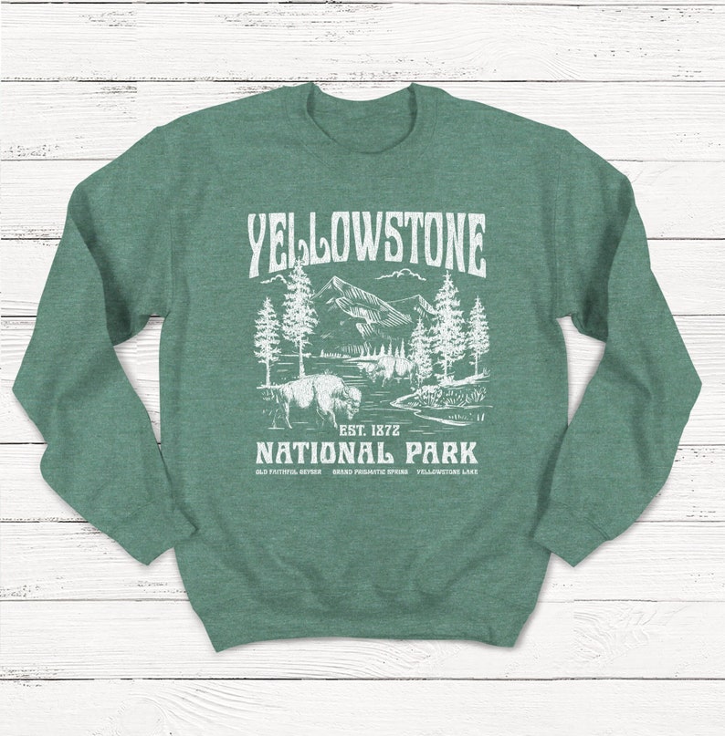 May include: Green sweatshirt with a white graphic design of a mountain landscape with trees and a bison. The text "Yellowstone National Park" is printed on the sweatshirt, along with "Est. 1872" and the names of some of the park's attractions.