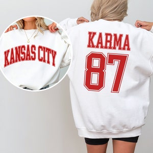 Op de afbeelding: Twee witte sweatshirts met rode letters. E&eacute;n sweatshirt zegt "Kansas City" en de andere zegt "Karma 87".
