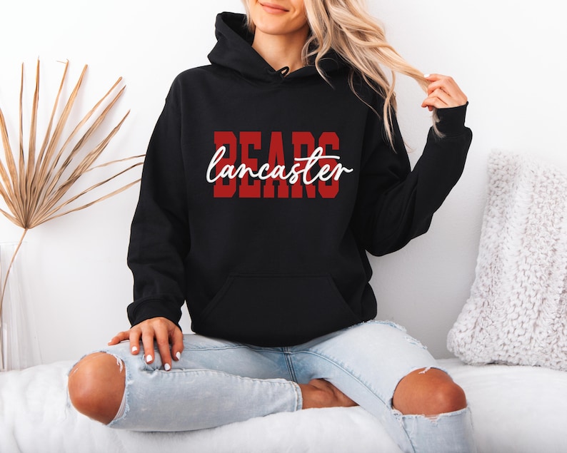 May include: Black hooded sweatshirt with the word "BEARS" in red block letters and "Lancaster" in white script. The sweatshirt has a front pocket and a drawstring hood. The model is wearing light blue jeans.