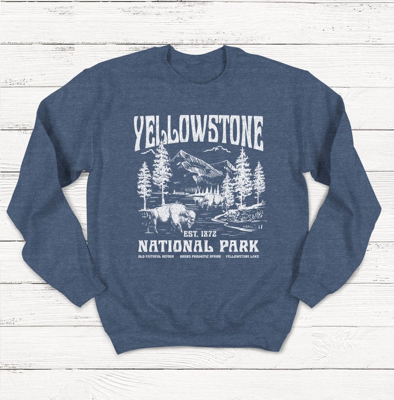 May include: A navy blue sweatshirt with a white graphic design of a mountain landscape, a bison, and the text "Yellowstone National Park Est. 1872 Old Faithful Geyser Grand Prismatic Spring Yellowstone Lake".