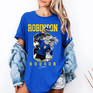 May include: Royal blue t-shirt featuring a volleyball player in action, with the text "ROBINSON HUDSON Lions" in yellow. The graphic includes a volleyball and the number 8. The shirt is a casual, sporty style.