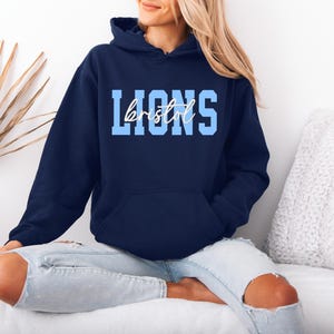 May include: A navy blue hooded sweatshirt with the word "LIONS" in light blue block letters and "bristol" in a cursive font. The sweatshirt has a front pocket and a drawstring hood. The model is wearing light blue jeans.