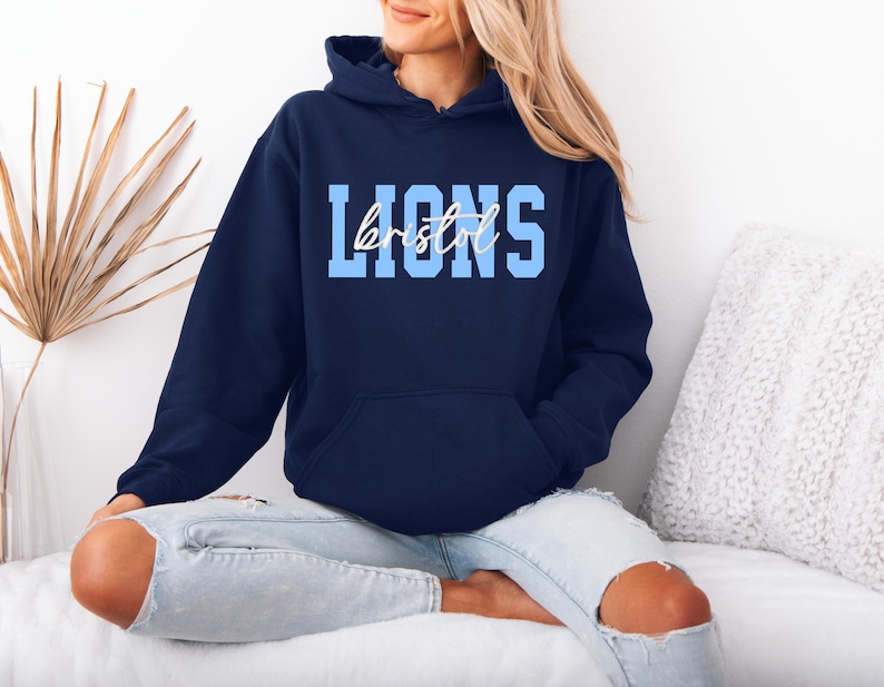 May include: A navy blue hooded sweatshirt with the word "LIONS" in light blue block letters and "bristol" in a cursive font. The sweatshirt has a front pocket and a drawstring hood. The model is wearing light blue jeans.