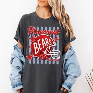 K&ouml;nnte beinhalten: Ein anthrazitfarbenes T-Shirt mit einer Grafik eines roten Football-Helms, dem wei&szlig;en Schriftzug BEARS, dem roten Wort Pinecrest dar&uuml;ber und dem Text Caleb #18 darunter. Das Design enth&auml;lt rote Sterne und einen hellblauen Hintergrund mit vertikalen Streifen.