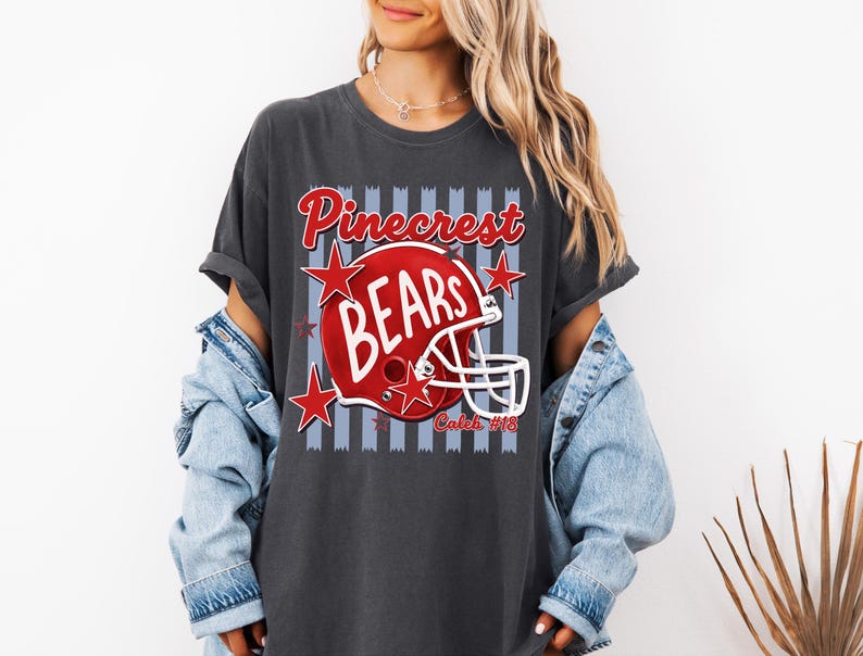 K&ouml;nnte beinhalten: Ein anthrazitfarbenes T-Shirt mit einer Grafik eines roten Football-Helms, dem wei&szlig;en Schriftzug BEARS, dem roten Wort Pinecrest dar&uuml;ber und dem Text Caleb #18 darunter. Das Design enth&auml;lt rote Sterne und einen hellblauen Hintergrund mit vertikalen Streifen.
