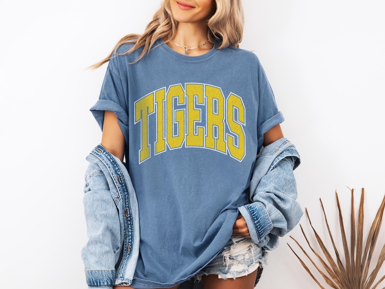 May include: Blue t-shirt with the word "TIGERS" in yellow block letters with a white outline. The shirt has rolled-up sleeves. A denim jacket is draped over the shoulders.