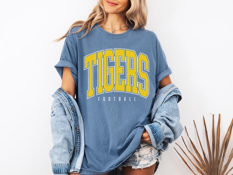 May include: A blue short-sleeve t-shirt featuring the word TIGERS in large yellow block letters with a white outline, positioned above the word FOOTBALL in smaller white text. The shirt has a crew neckline and a relaxed, loose-fitting silhouette.