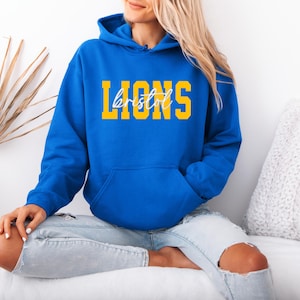 May include: A royal blue hoodie with a front pocket and a drawstring hood. The word "LIONS" is printed in yellow block letters, with "bristol" in a cursive font underneath. The hoodie is worn with light blue jeans.