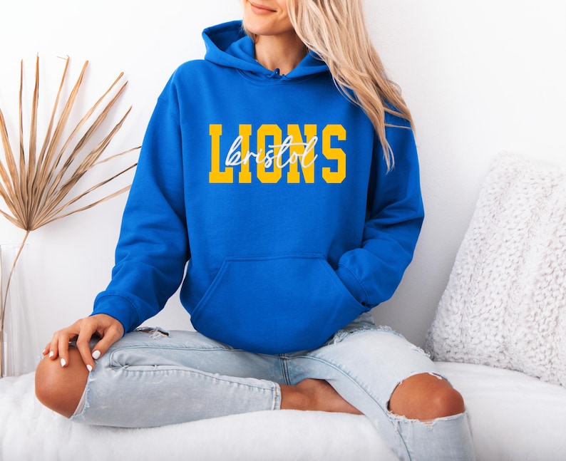 May include: A royal blue hoodie with a front pocket and a drawstring hood. The word "LIONS" is printed in yellow block letters, with "bristol" in a cursive font underneath. The hoodie is worn with light blue jeans.