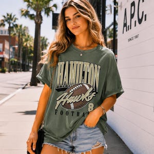 Puede incluir: Una camiseta de manga corta color verde oliva con un estampado gr&aacute;fico que incluye el texto HAMILTON HAWKS FOOTBALL y una ilustraci&oacute;n de un bal&oacute;n de f&uacute;tbol americano marr&oacute;n. La prenda tiene cuello redondo y un corte holgado con mangas vueltas.