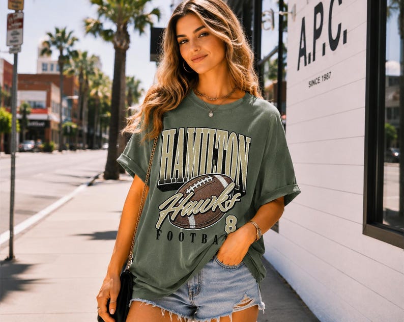 Puede incluir: Una camiseta de manga corta color verde oliva con un estampado gr&aacute;fico que incluye el texto HAMILTON HAWKS FOOTBALL y una ilustraci&oacute;n de un bal&oacute;n de f&uacute;tbol americano marr&oacute;n. La prenda tiene cuello redondo y un corte holgado con mangas vueltas.