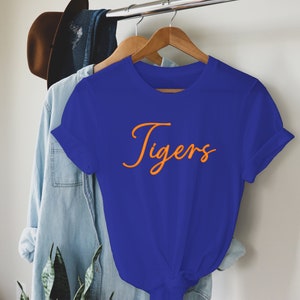 May include: A royal blue t-shirt with short sleeves and a crew neck. The shirt has the word "Tigers" written in orange cursive lettering.