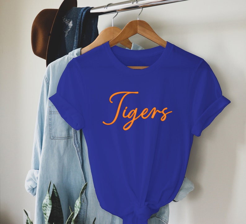 May include: A royal blue t-shirt with short sleeves and a crew neck. The shirt has the word "Tigers" written in orange cursive lettering.