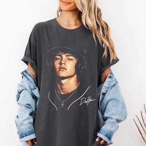 May include: Dark gray t-shirt featuring a portrait of a young man wearing a hat and headphones. The image includes the name "Dalton" in a cursive script. The shirt is paired with a light blue denim jacket.