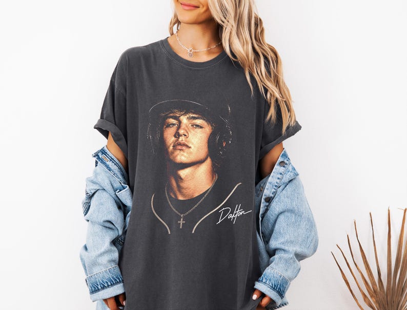 May include: Dark gray t-shirt featuring a portrait of a young man wearing a hat and headphones. The image includes the name "Dalton" in a cursive script. The shirt is paired with a light blue denim jacket.