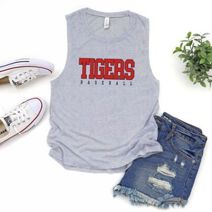 May include: Gray sleeveless tank top with the word "TIGERS" in red and "BASEBALL" in blue underneath. The tank top is laying on a white surface with a pair of white sneakers and denim shorts.