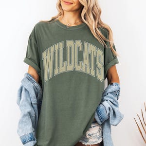 May include: Olive green t-shirt with the word "WILDCATS" in a beige, arched, collegiate-style font. The shirt has rolled-up sleeves and is worn with a denim jacket. The overall style is casual and relaxed.