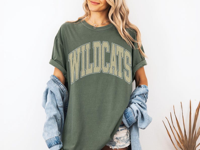 May include: Olive green t-shirt with the word "WILDCATS" in a beige, arched, collegiate-style font. The shirt has rolled-up sleeves and is worn with a denim jacket. The overall style is casual and relaxed.