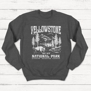 May include: Gray sweatshirt with a white graphic design of a mountain landscape with trees and a bison. The text "Yellowstone National Park" is printed below the image, along with "Est. 1872" and "Old Faithful Geyser, Grand Prismatic Spring, Yellowstone Lake."