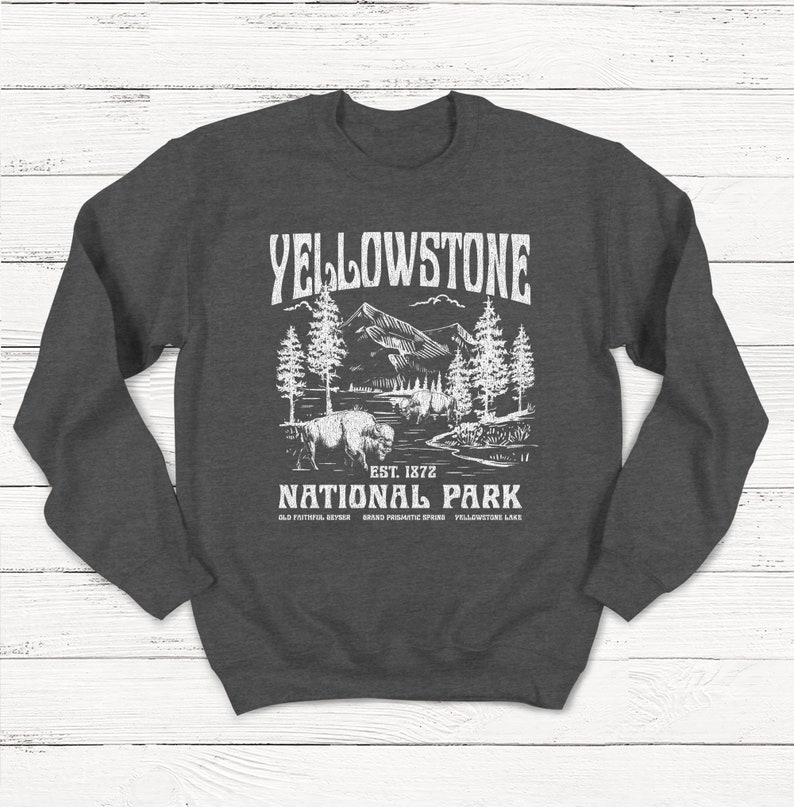 May include: Gray sweatshirt with a white graphic design of a mountain landscape with trees and a bison. The text "Yellowstone National Park" is printed below the image, along with "Est. 1872" and "Old Faithful Geyser, Grand Prismatic Spring, Yellowstone Lake."