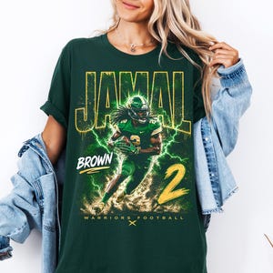 May include: Dark green t-shirt featuring a football player graphic with the name "JAMAL" and "BROWN" in yellow. The number "2" is also in yellow. The words "WARRIORS FOOTBALL" are at the bottom.