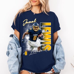 K&ouml;nnte beinhalten: Ein marineblaues Kurzarm-T-Shirt mit einem Grafikdruck eines Footballspielers in einer marine-goldenen Uniform mit der Nummer 8. Der Text lautet Jamal Lewis, Lions und die Nummer 8. Das Design enth&auml;lt Blitzeffekte und eine kr&auml;ftige gelbe Schrift.