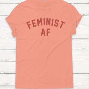 May include: A peach colored t-shirt with the text "FEMINIST AF" in a red color.