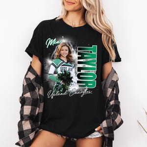 May include: Black t-shirt featuring a photo of a cheerleader in a green and white uniform, with the name "Taylor" in green vertical text. The shirt also includes the words "Mia" and "Upland Chargers".