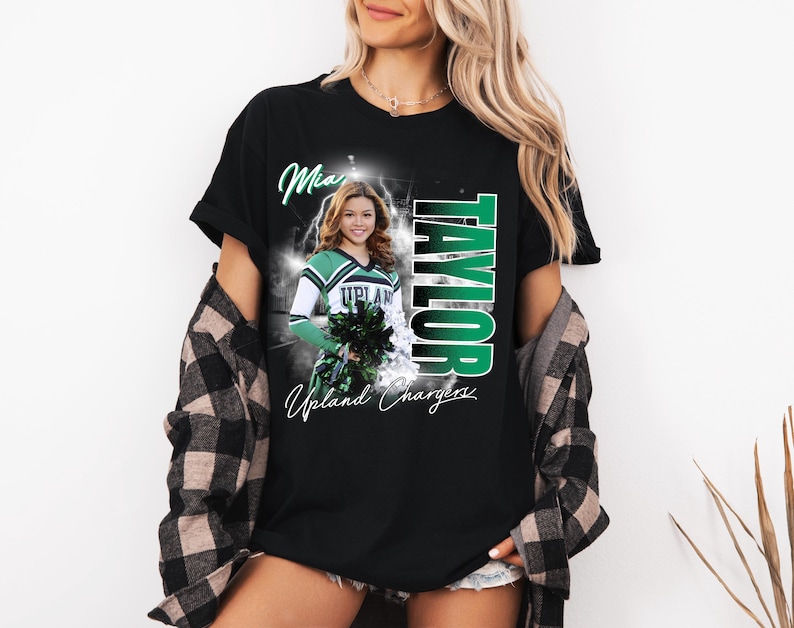 May include: Black t-shirt featuring a photo of a cheerleader in a green and white uniform, with the name "Taylor" in green vertical text. The shirt also includes the words "Mia" and "Upland Chargers".