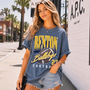 Puede incluir: Una camiseta de manga corta color azul pizarra con un estampado gr&aacute;fico que incluye el texto BENTON en amarillo, Bulldogs en letra cursiva amarilla y FOOTBALL en blanco, junto a una ilustraci&oacute;n de un bal&oacute;n de f&uacute;tbol americano marr&oacute;n y el n&uacute;mero 23 en amarillo.