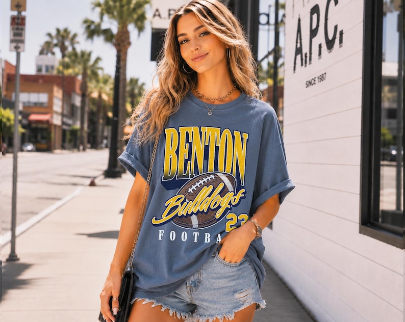 Puede incluir: Una camiseta de manga corta color azul pizarra con un estampado gr&aacute;fico que incluye el texto BENTON en amarillo, Bulldogs en letra cursiva amarilla y FOOTBALL en blanco, junto a una ilustraci&oacute;n de un bal&oacute;n de f&uacute;tbol americano marr&oacute;n y el n&uacute;mero 23 en amarillo.