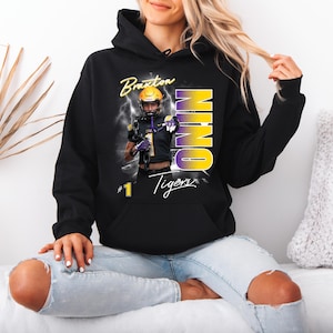 May include: Black hooded sweatshirt featuring a football player graphic with the name "Braxton" and the word "NINO" in yellow and purple. The sweatshirt also has the text "Tigers" and the number "1".