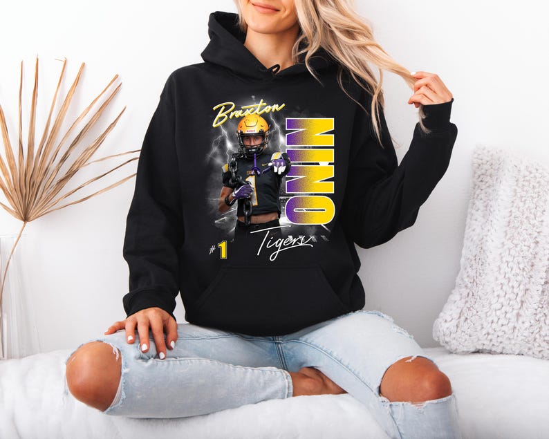 May include: Black hooded sweatshirt featuring a football player graphic with the name "Braxton" and the word "NINO" in yellow and purple. The sweatshirt also has the text "Tigers" and the number "1".