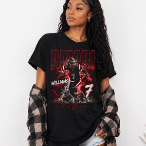 May include: Black t-shirt featuring a football player graphic with the name "AMARI" in red, along with "WILLIAMS" and the number "7". The design includes a red lightning effect and the words "TIGERS FOOTBALL".