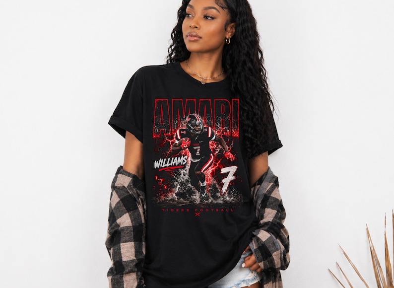 May include: Black t-shirt featuring a football player graphic with the name "AMARI" in red, along with "WILLIAMS" and the number "7". The design includes a red lightning effect and the words "TIGERS FOOTBALL".
