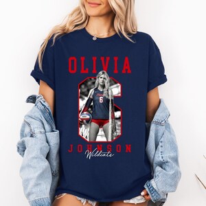 May include: Navy blue t-shirt featuring a volleyball player's image, the name "OLIVIA JOHNSON" in red, and the word "Wildcats" in white script. The player is wearing a red and navy uniform with the number 6.