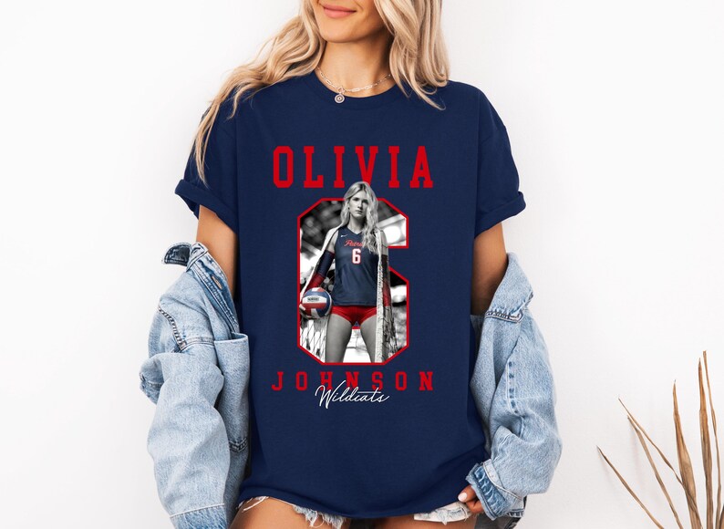 May include: Navy blue t-shirt featuring a volleyball player's image, the name "OLIVIA JOHNSON" in red, and the word "Wildcats" in white script. The player is wearing a red and navy uniform with the number 6.