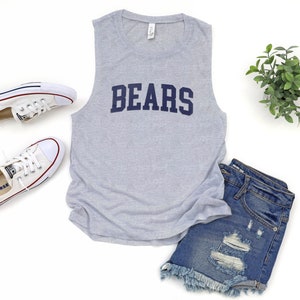 May include: A light gray sleeveless shirt with the word "BEARS" in navy blue block letters. The shirt is paired with denim shorts and white sneakers. A small green plant is also in the image.
