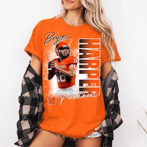 K&ouml;nnte beinhalten: Ein orangefarbenes T-Shirt mit einem Grafikdruck eines Footballspielers im orangefarbenen Trikot mit der Nummer 16, dem Text Bryce Harper und dem Wort Panthers. Das T-Shirt wird unter einem schwarz-wei&szlig; karierten Langarmhemd mit Kn&ouml;pfen getragen.