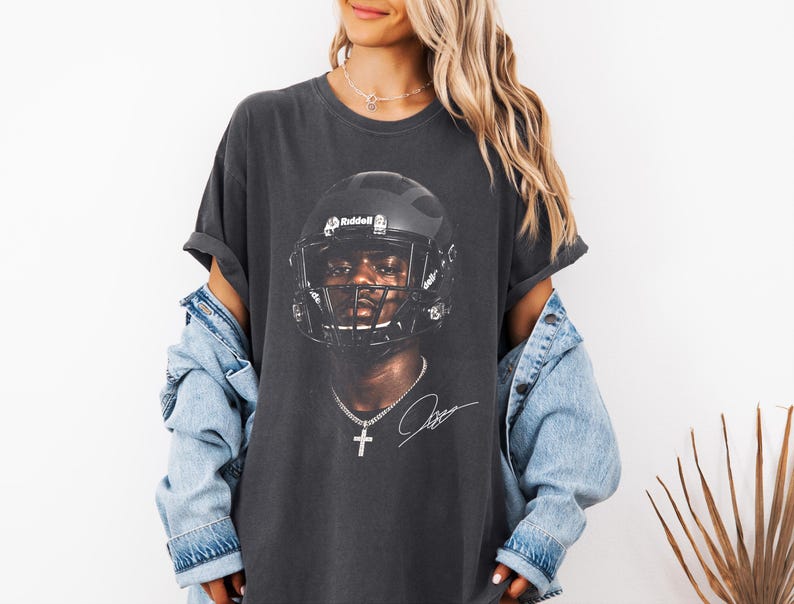 May include: Dark gray t-shirt featuring a football player wearing a black helmet. The image includes a signature and a cross pendant. The shirt is worn with a light blue denim jacket.
