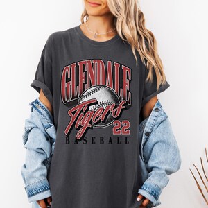May include: A charcoal gray short-sleeve t-shirt featuring a graphic print with the text GLENDALE TIGERS 22 BASEBALL and an illustration of a baseball. The shirt is worn with a light blue denim jacket layered over the shoulders.