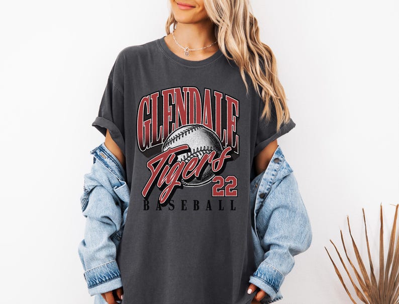 May include: A charcoal gray short-sleeve t-shirt featuring a graphic print with the text GLENDALE TIGERS 22 BASEBALL and an illustration of a baseball. The shirt is worn with a light blue denim jacket layered over the shoulders.