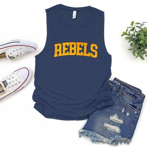 May include: Navy blue sleeveless shirt with the word "REBELS" in gold lettering. The shirt is paired with denim shorts and white sneakers. A sprig of greenery adds a decorative touch to the scene.
