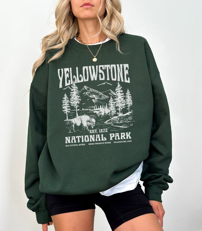 May include: A dark green sweatshirt with a white graphic print of a mountain scene and the text "Yellowstone National Park" with the words "Est. 1872" and "Old Faithful Geyser, Grand Prismatic Spring, Yellowstone Lake" underneath.