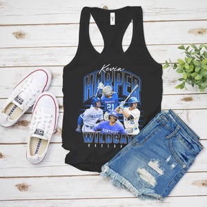 K&ouml;nnte beinhalten: Ein schwarzes Racerback-Tanktop mit einem Grafikdruck von Baseballspielern in blau-wei&szlig;en Uniformen. Der Text lautet Kevin Harper und Kentucky Wildcats Baseball. Das Design zeigt mehrere Action-Posen der Spieler auf dem Spielfeld.