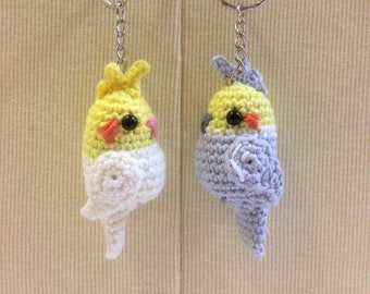 Può includere: Due portachiavi a forma di cacatua crochet, uno giallo e uno grigio, con occhi neri e becchi arancioni. I portachiavi sono attaccati a anelli portachiavi argentati.