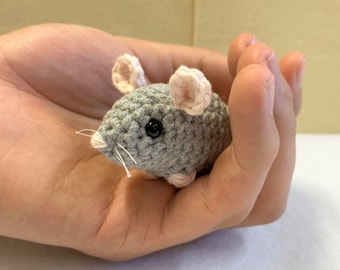 Può includere: Un piccolo topo di uncinetto grigio con orecchie rosa. Il topo è tenuto nella mano di una persona.