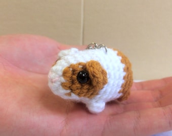 Può includere: Un piccolo portachiavi a forma di cavia fatto a crochet con pelliccia bianca e marrone, occhi neri e naso rosa.