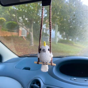 Pu&ograve; includere: Un ornamento all'uncinetto a forma di cacatua bianco con cresta gialla, occhi a perla neri e becco grigio, appollaiato su un piccolo bastoncino di legno. L'uccello &egrave; sospeso da due fili di lana marrone, per essere appeso allo specchietto.