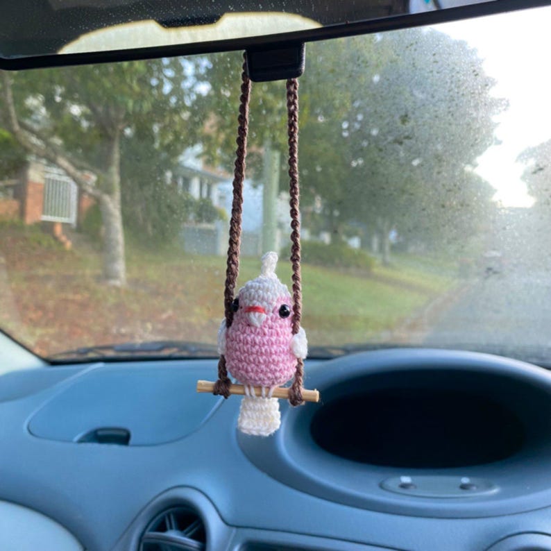 Pu&ograve; includere: Un ornamento a forma di uccellino lavorato all'uncinetto in rosa e bianco, appeso a un cordino di filo marrone. L'uccellino presenta una piccola cresta, occhi a perla neri e un becco arancione chiaro, appollaiato su un bastoncino di legno.